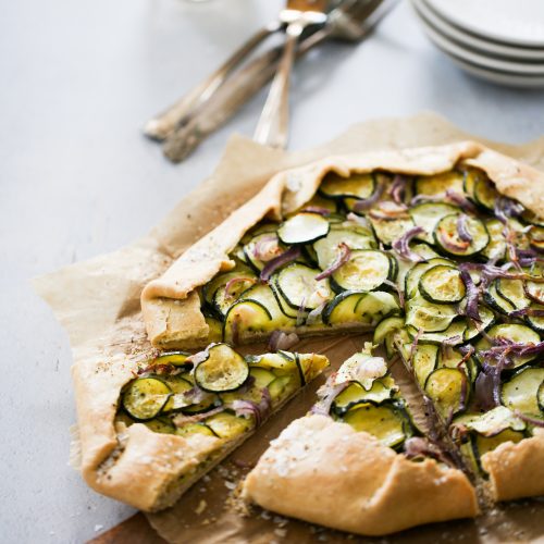A rustic zucchini galette topped with sliced zucchini and red onions on parchment paper. The galette has one slice cut and slightly removed from the whole. Behind the galette are stacked plates, forks, and a glass. The scene is set on a light-colored surface, creating a cozy and inviting atmosphere.