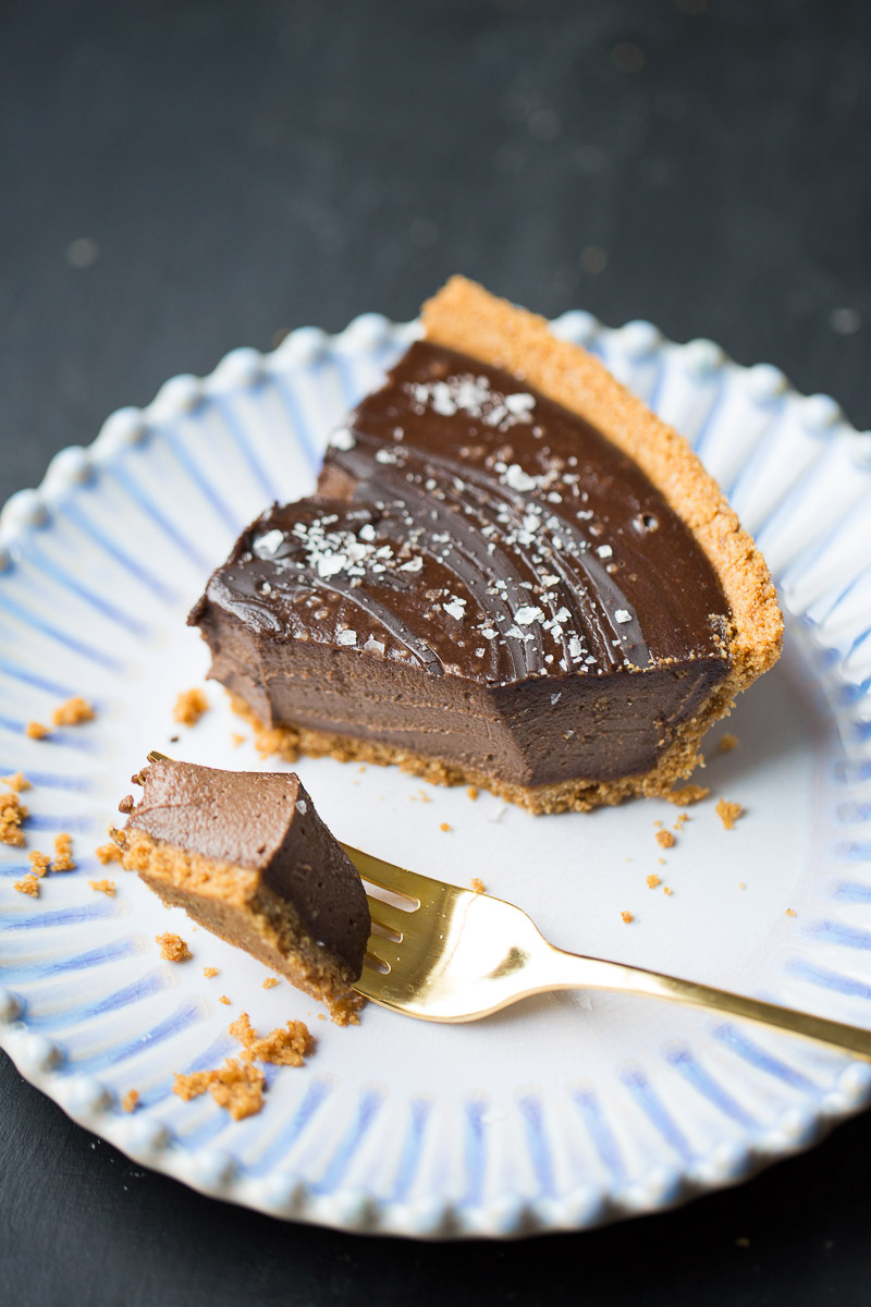 A slice of creamy chocolate tart with a graham cracker crust on a white patterned plate with a gold fork taking a bite out of it, garnished with a sprinkle of sea salt on top