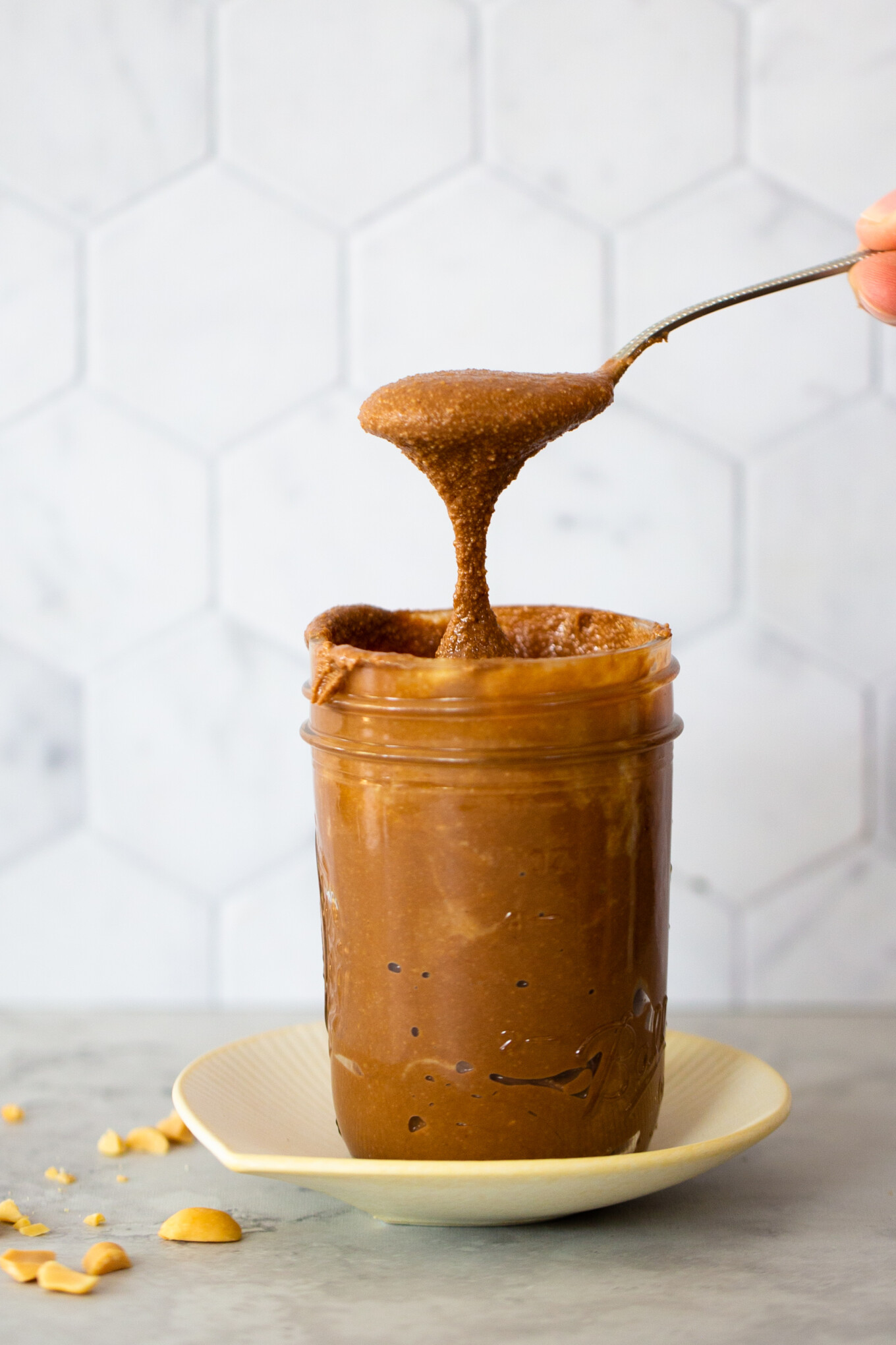 A spoon lifts a dollop of gooey, dark chocolate peanut butter from a full mason jar set on a small cream-colored plate. Peanut pieces are scattered on the gray countertop. The background features a white hexagonal tile pattern.