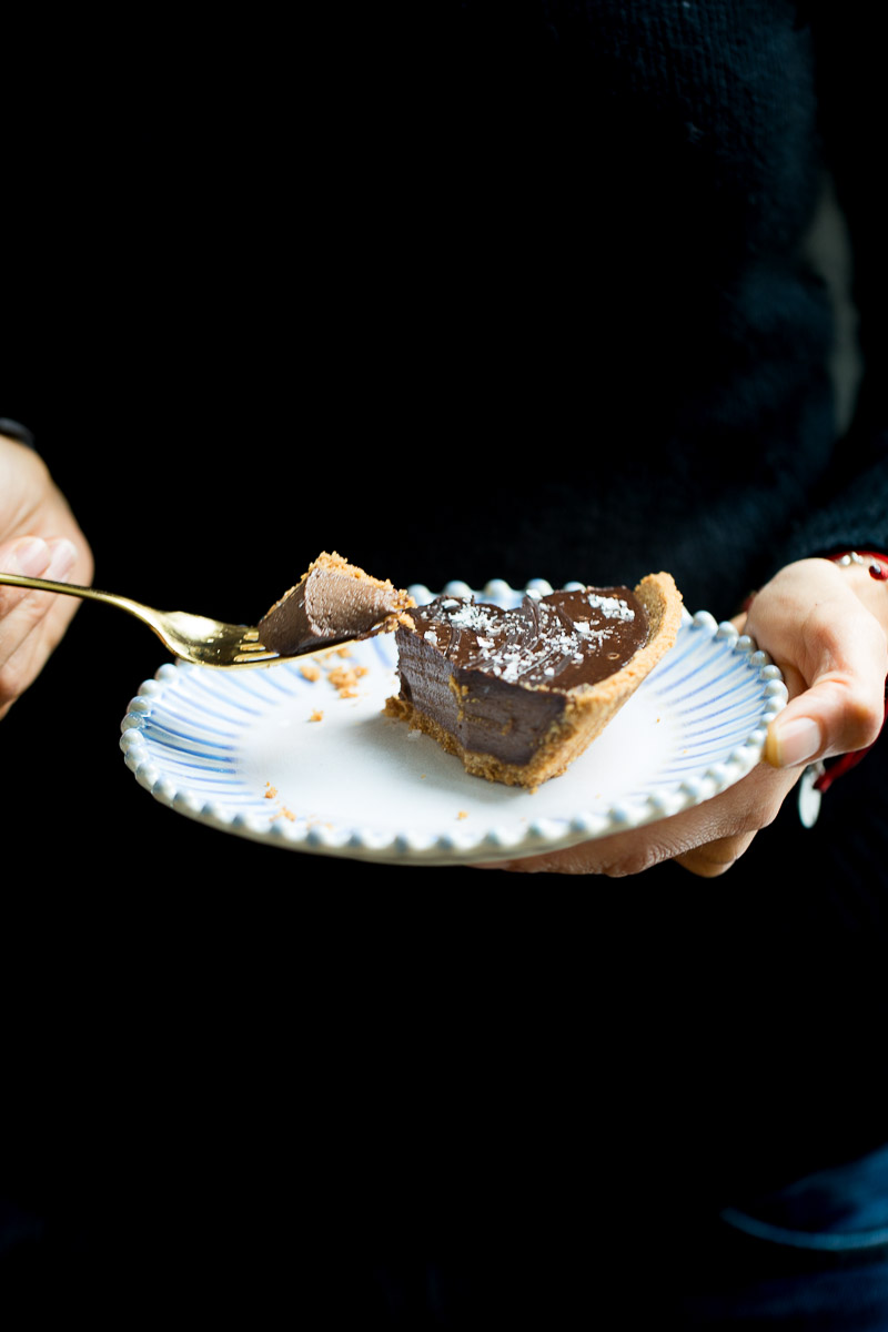 A person holding a plate with a slice of creamy chocolate vegan pie, taking a bite with a fork. The pie is garnished with powdered sugar and the background is dark.