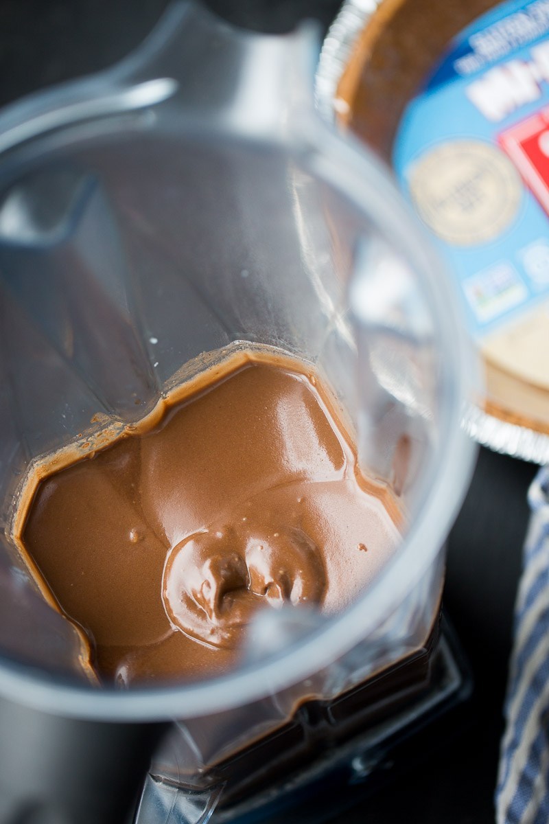 Close-up of a blender jug containing freshly blended creamy chocolate vegan pie filling, with the texture appearing creamy and smooth, shown in-focus with a blurred background of a kitchen setting.