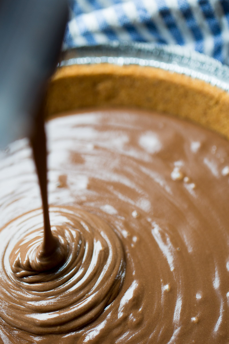 Creamy vegan chocolate is being poured into a pie crust, creating a spiral pattern. The pie is on a blue and white striped cloth, with a focus on the smooth, shiny texture of the chocolate
