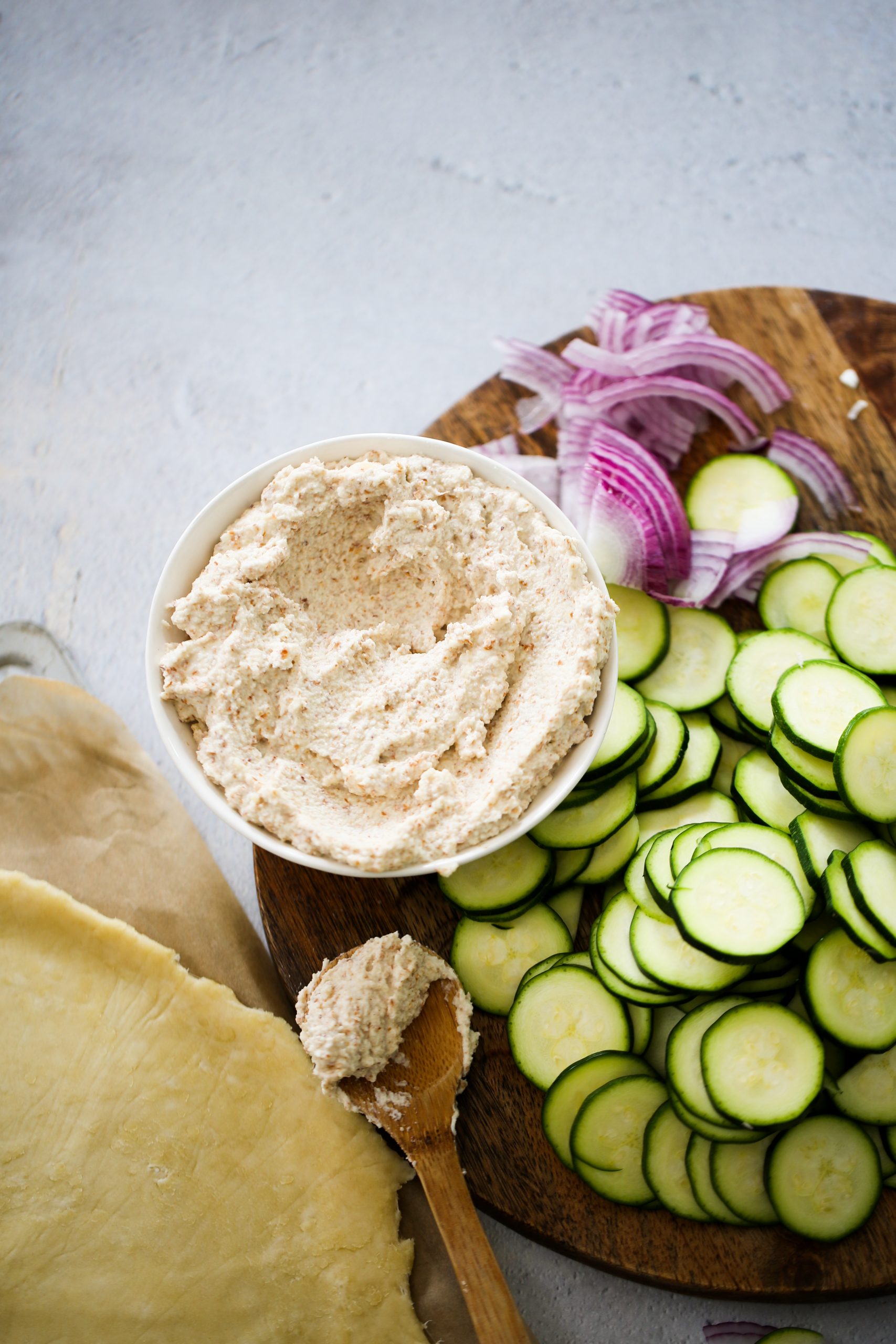 A bowl of creamy ricotta cheese on a wooden platter is surrounded by thinly sliced zucchini and red onion, perfect for assembling a savory zucchini galette. A wooden spoon with a small amount of cheese rests nearby on a light, textured surface. A rolled-out dough lies next to the platter, partially visible on parchment paper.