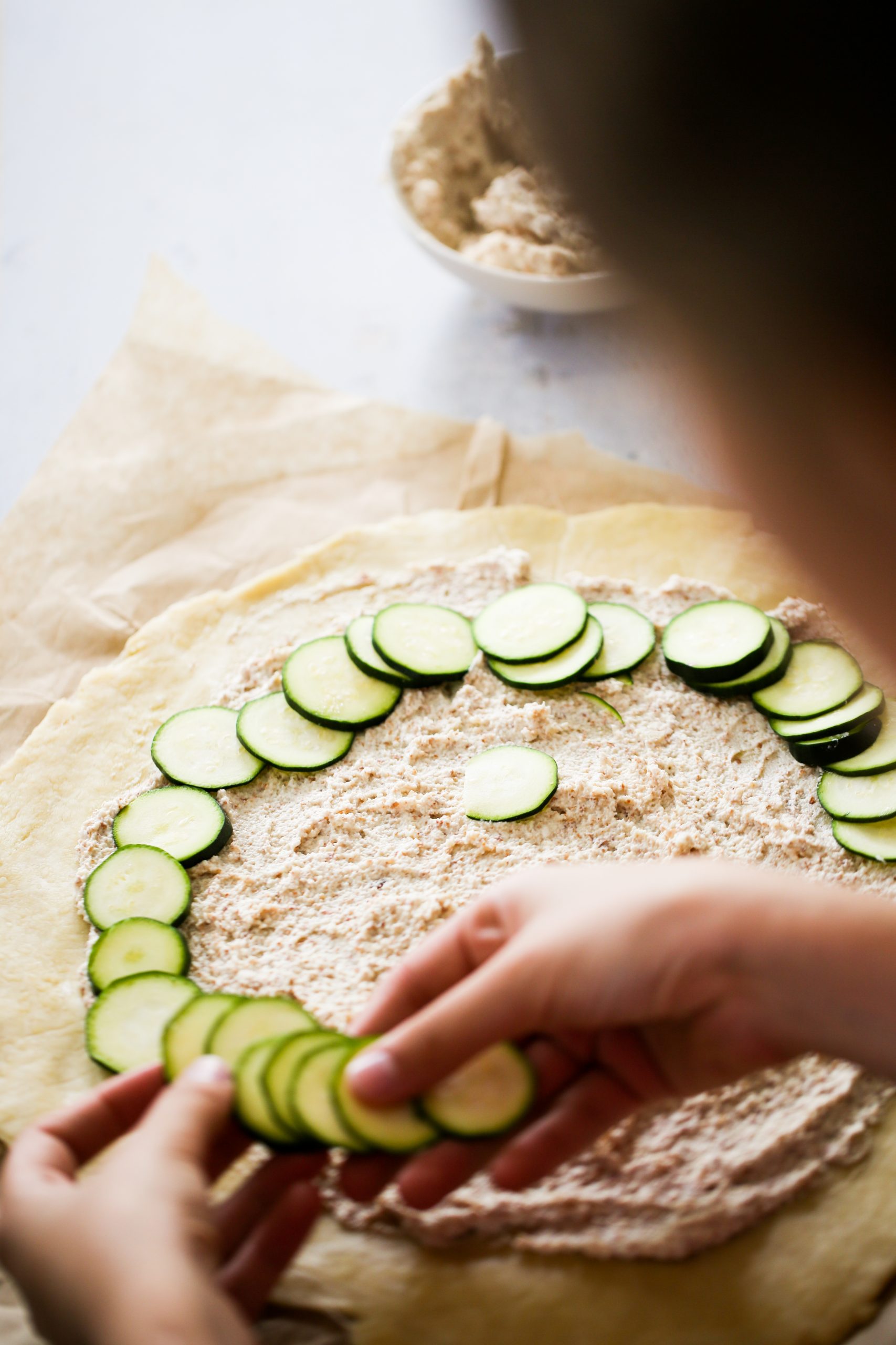 A person is arranging slices of cucumber in a circular pattern on dough spread with a white creamy mixture, creating a delicate zucchini galette. The dough rests on parchment paper, and in the background, slightly out of focus, sits a bowl filled with more of the creamy mixture.
