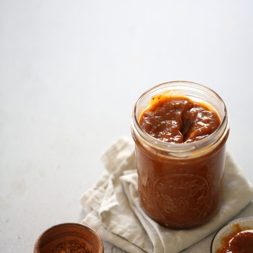 A jar of homemade chamoy sauce, partially filled, on a white cloth, with a wooden spoon and a small bowl of spices beside it, all set on a light gray surface. The ambiance is