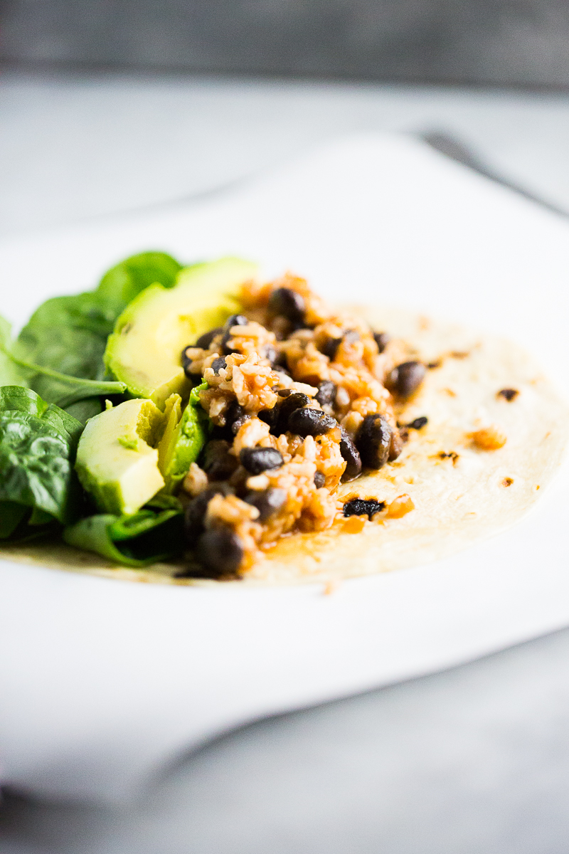 A close-up of a soft tortilla, perfect for burritos, topped with black beans, rice, avocado slices, and fresh spinach. The tortilla rests on a white surface, and the background is slightly blurred, highlighting the vibrant ingredients. The light creates a bright and clean aesthetic.