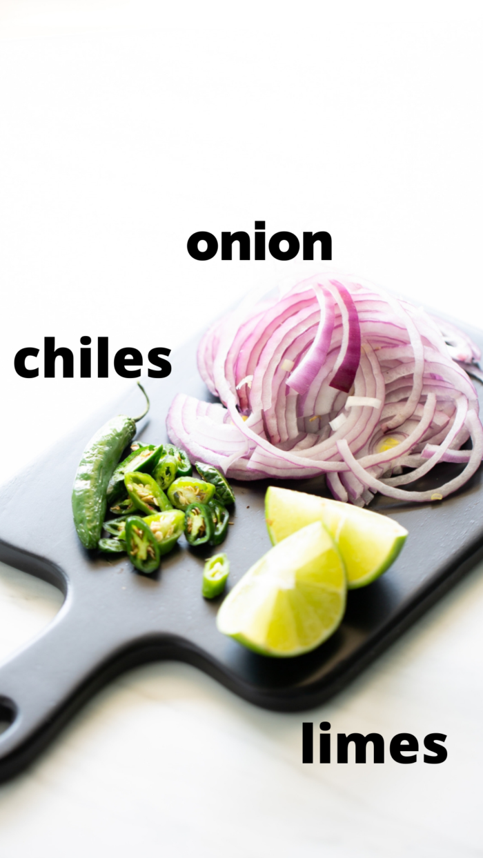 A black cutting board with chopped ingredients on a white background. It features sliced red onions, chopped green chiles toreados, and quartered limes. Text labels identify each ingredient: "onion" above the slices, "chiles toreados" to the left of the pieces, and "limes" above the lime wedges.
