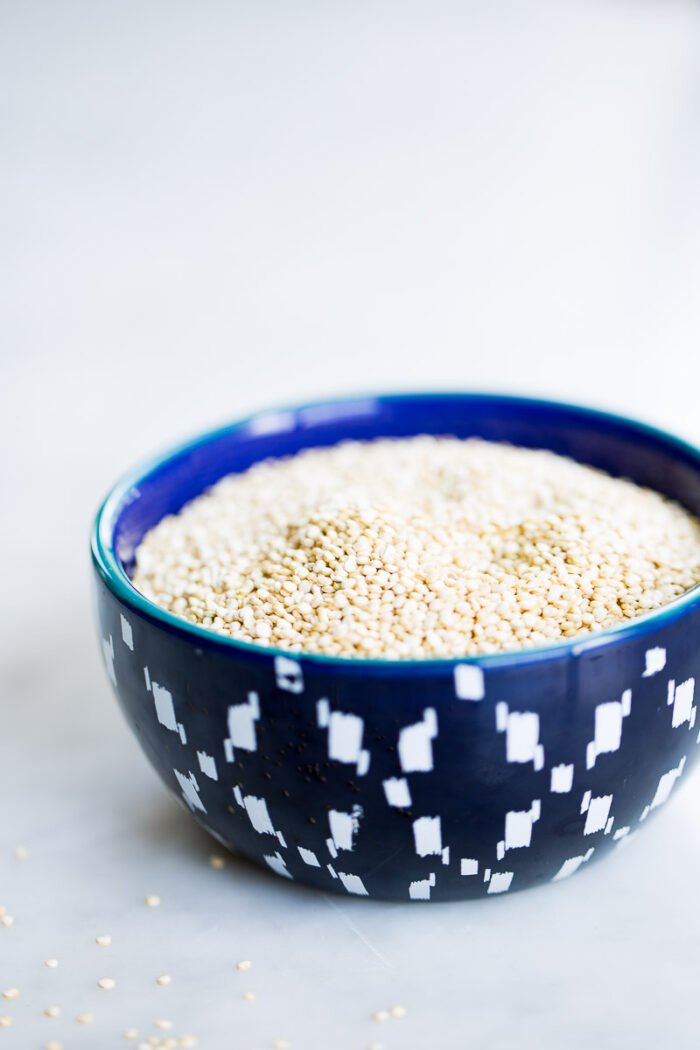 A close-up shot of a dark blue ceramic bowl with an abstract white pattern. The bowl is filled with uncooked white quinoa seeds, perfect for creating a refreshing quinoa summer salad. A few seeds are scattered on the white surface it rests on, while the soft-focused background emphasizes the texture and design.