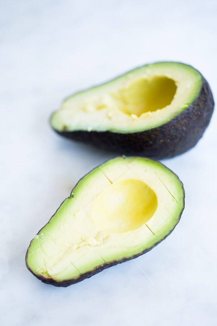 A ripe avocado cut in half with the pit removed. The halves are placed on a white surface; the front half has scored lines in the flesh. Perfect for a quinoa summer salad, the skin is dark green and rough, while the flesh is a light green color.