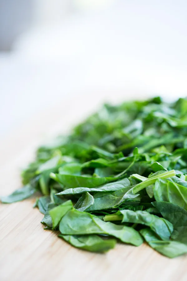 A close-up shot of a pile of fresh spinach leaves spread on a light wooden surface. The green leaves appear crisp and vibrant, with bright light illuminating them from above, creating a fresh and appetizing look—perfect for tossing into a quinoa summer salad. The background is softly blurred, keeping the focus on the spinach.