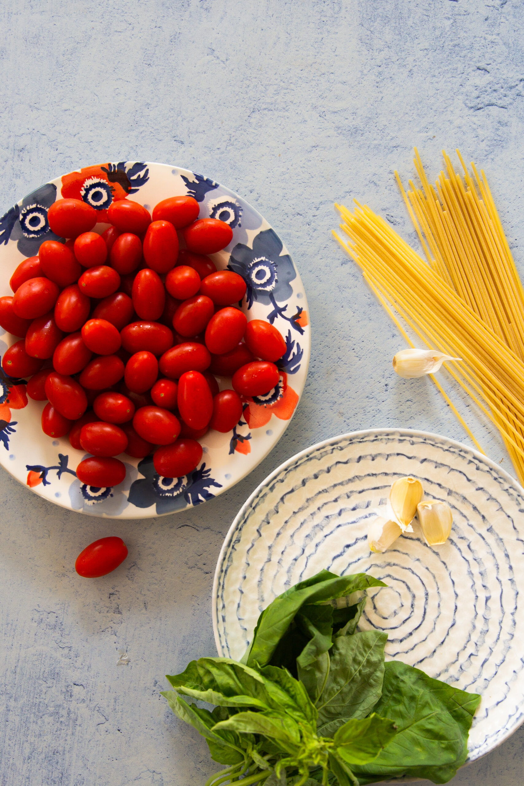 Plate of cherry tomatoes, raw spaghetti, garlic cloves and fresh basil on a light blue surface for pasta with cherry tomatoes and basil.