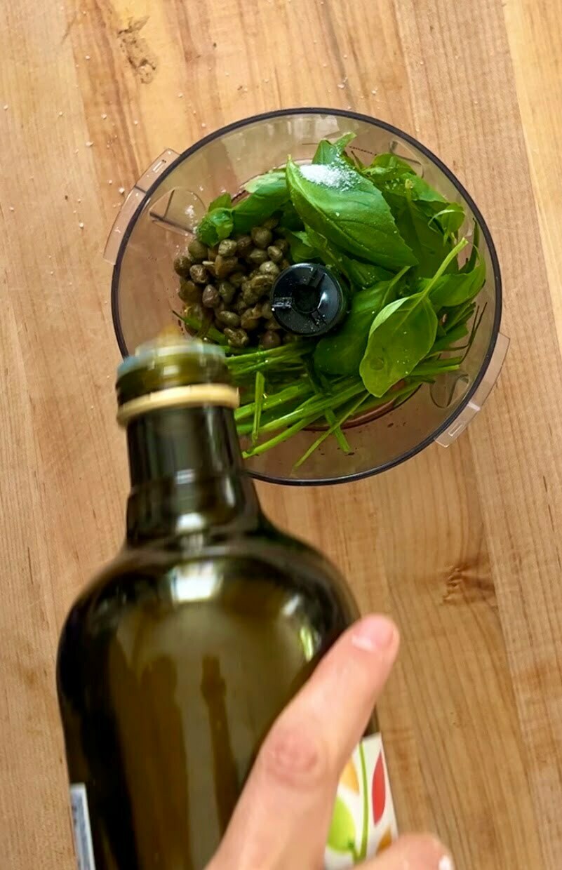 Una mano vierte aceite de oliva de una botella verde en el recipiente de un procesador de alimentos. Dentro del recipiente hay hojas de albahaca verde fresca y alcaparras, ingredientes perfectos para una receta. El procesador de alimentos está colocado sobre una encimera de madera, rodeado de verduras de colores vivos. La imagen está tomada desde arriba.