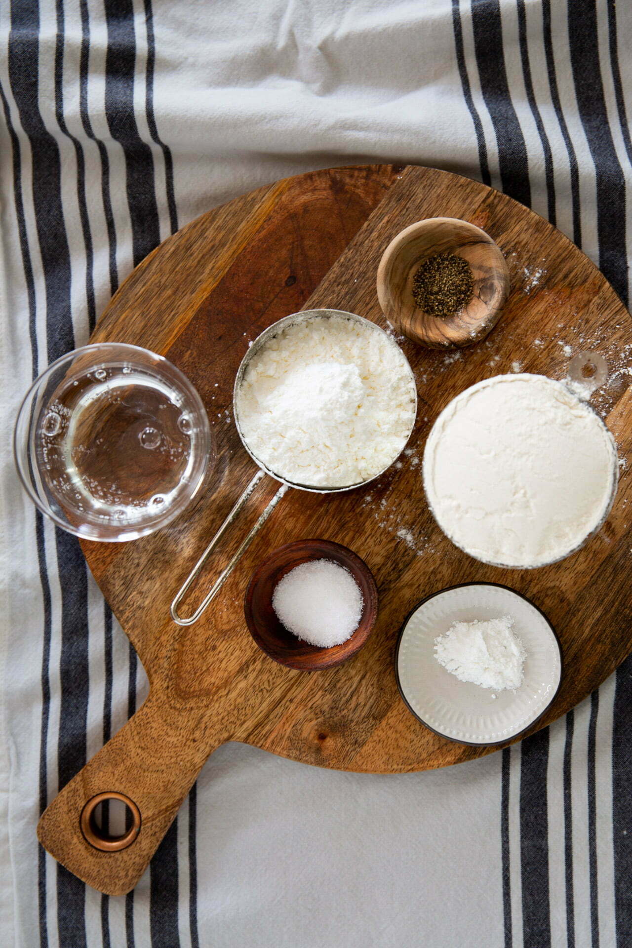 Una tabla de cortar de madera contiene varios ingredientes para hornear sobre un paño rayado, que recuerda a la preparación de una receta italiana. Entre los elementos se incluyen un vaso de agua, una taza de metal con harina, un cuenco pequeño con semillas de amapola, un cuenco pequeño con sal y un plato con una sustancia blanca en polvo.