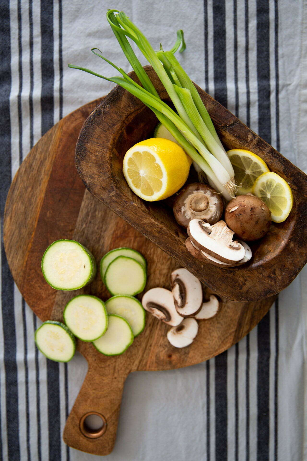 Una tabla de cortar de madera muestra rodajas de calabacín y champiñones junto a un cuenco de madera rectangular que contiene cebollas verdes, un limón entero, rodajas de limón y champiñones adicionales: todas las verduras frescas necesarias para una deliciosa receta de frito misto. Los elementos descansan sobre un mantel a rayas.