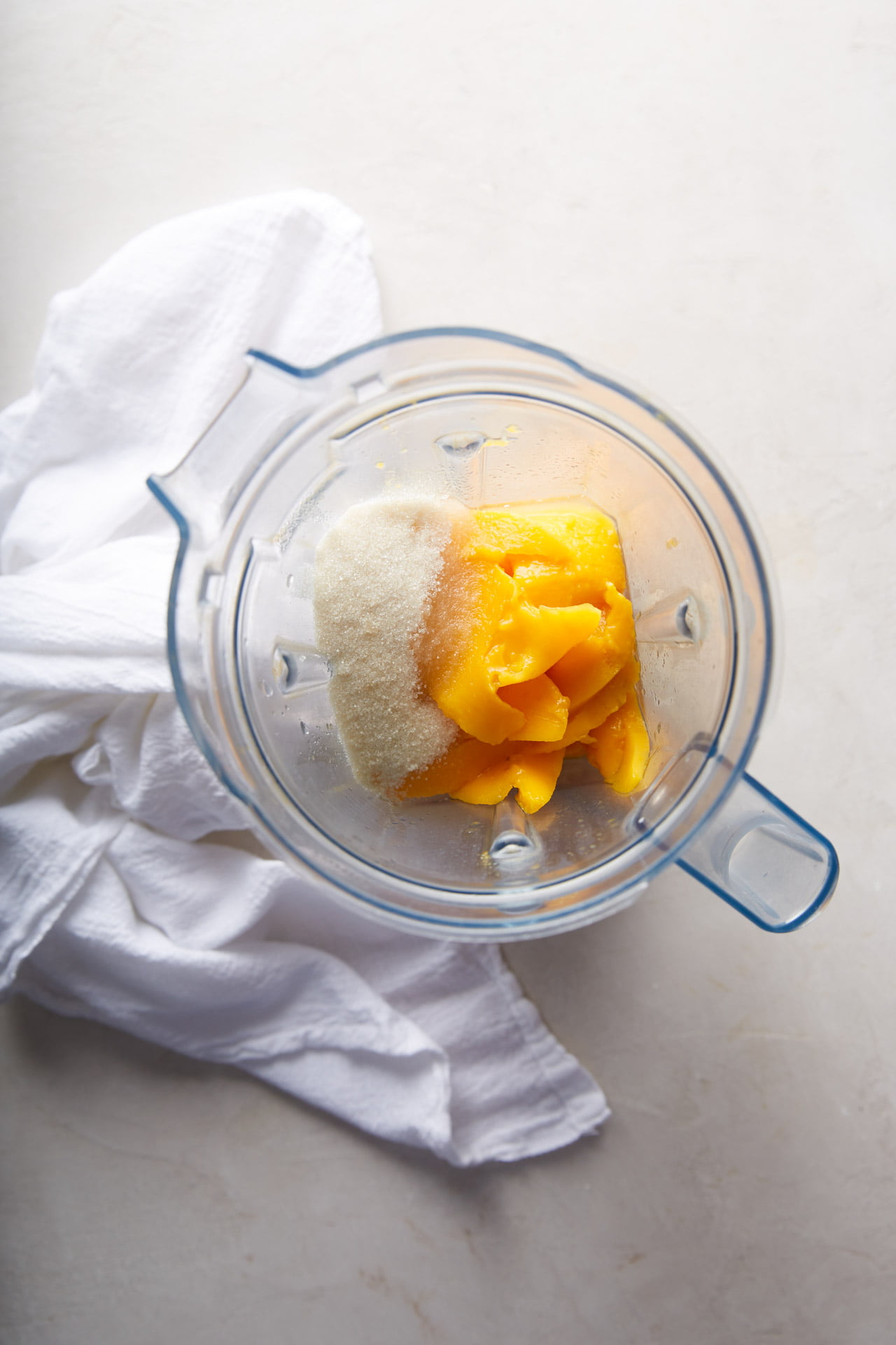 A top view of a blender containing mango slices and sugar, perfect for making Mexican mango drink, placed on a light-colored countertop. Beside the blender, there is a white cloth neatly arranged. The transparent blender allows a clear view of the ingredients inside. The overall setting appears bright and clean.