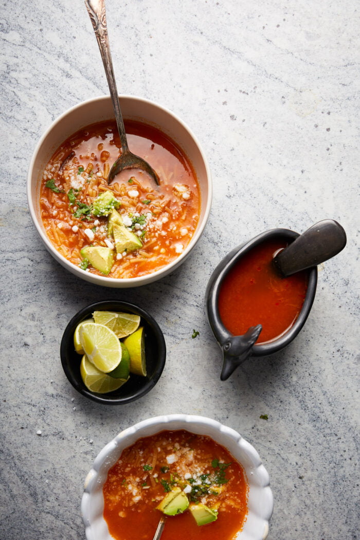 Two bowls of fideo soup garnished with avocado, cheese, and herbs. Nearby are lime wedges in a small black bowl and a dolphin-shaped sauce boat with additional soup. Each bowl has a spoon. The background is light gray, adding contrast to the vibrant tomato soup.