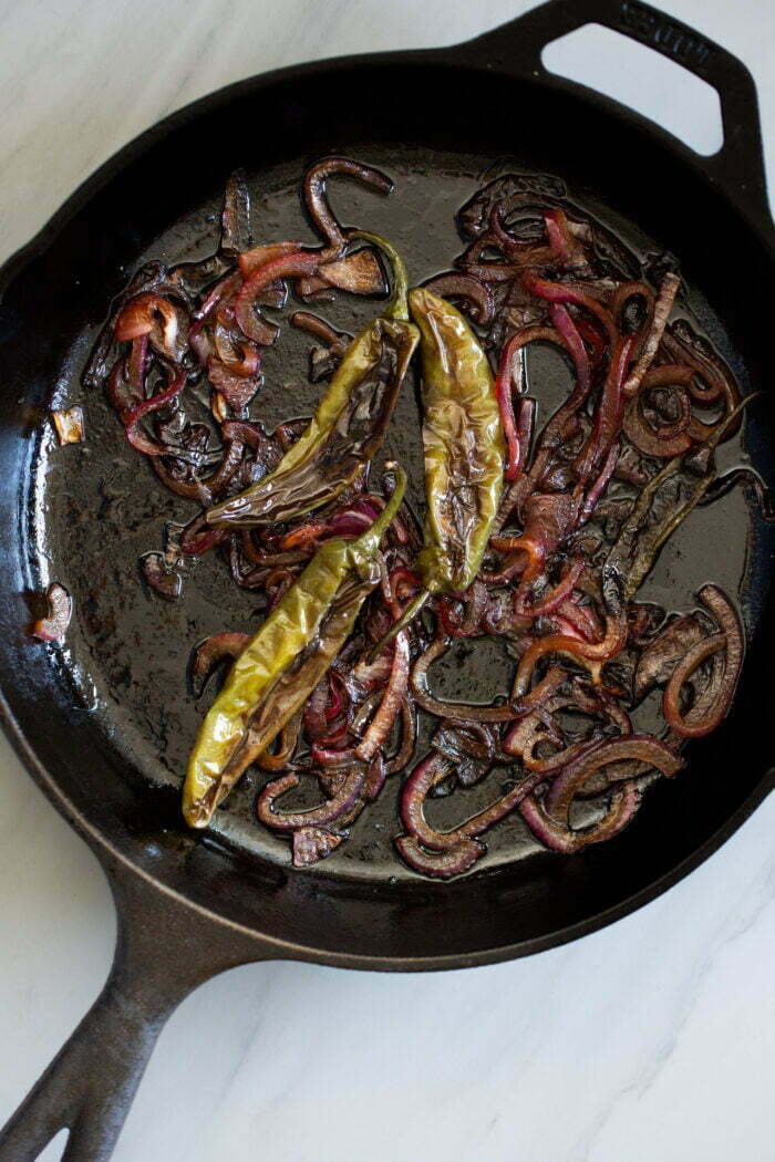 A cast-iron skillet contains charred vegetables, including several green peppers, caramelized red onion slices, and chiles toreados. The sautéed and slightly burnt vegetables add a rustic look to the dish. The skillet sits on a light-colored marble surface.