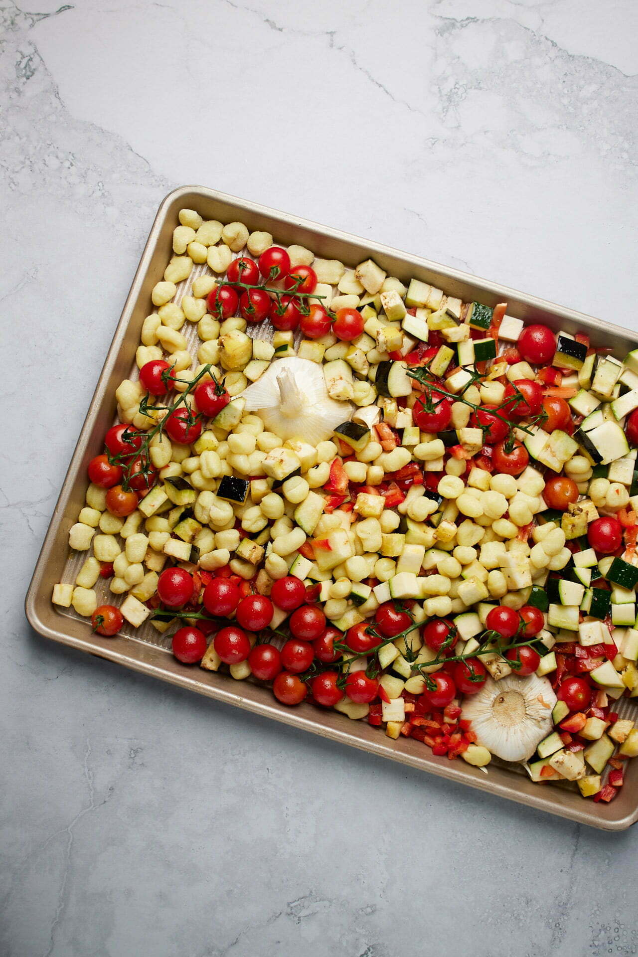 A sheet pan filled with chopped vegetables, including yellow potatoes, zucchini, red bell peppers, cherry tomatoes, and whole garlic cloves, sits on a light marble surface. The vibrant array of colors is beautifully complemented by dollops of herbed butter throughout the vegetables before roasting.