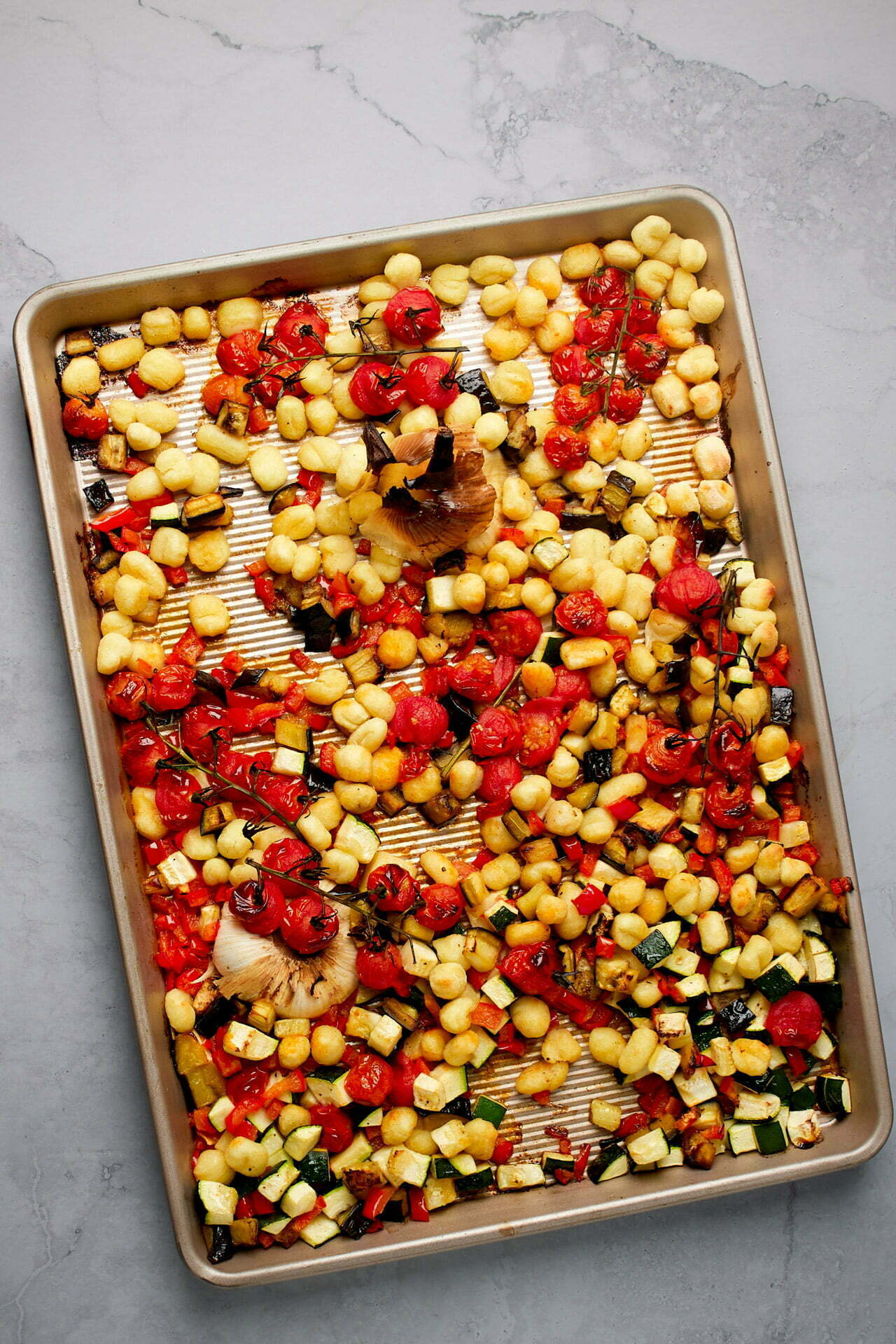 A baking sheet filled with roasted vegetables, including cherry tomatoes, zucchini, and onions, alongside golden brown sheet pan gnocchi, all spread evenly across. The vegetables are charred at the edges, creating a colorful and appetizing mixture drizzled with herbed butter. The background is a light gray, stone-textured surface.