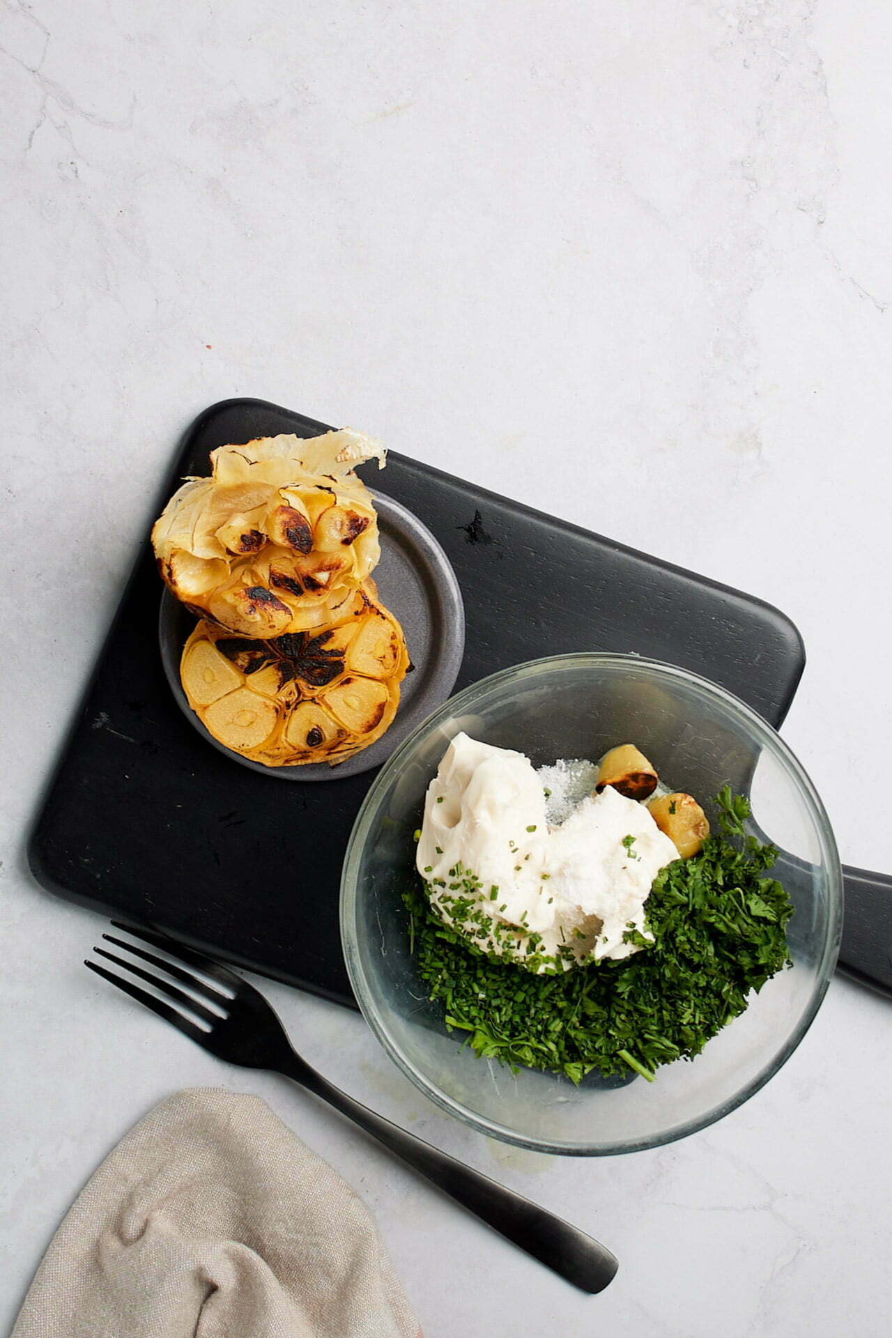 Overhead view of ingredients on a black cutting board and a white surface. A glass bowl contains a mixture of chopped green herbs, what appears to be sour cream, and roasted garlic. To the left, there's herbed butter next to a halved bulb of roasted garlic. A fork and a beige napkin are placed in the bottom left corner.