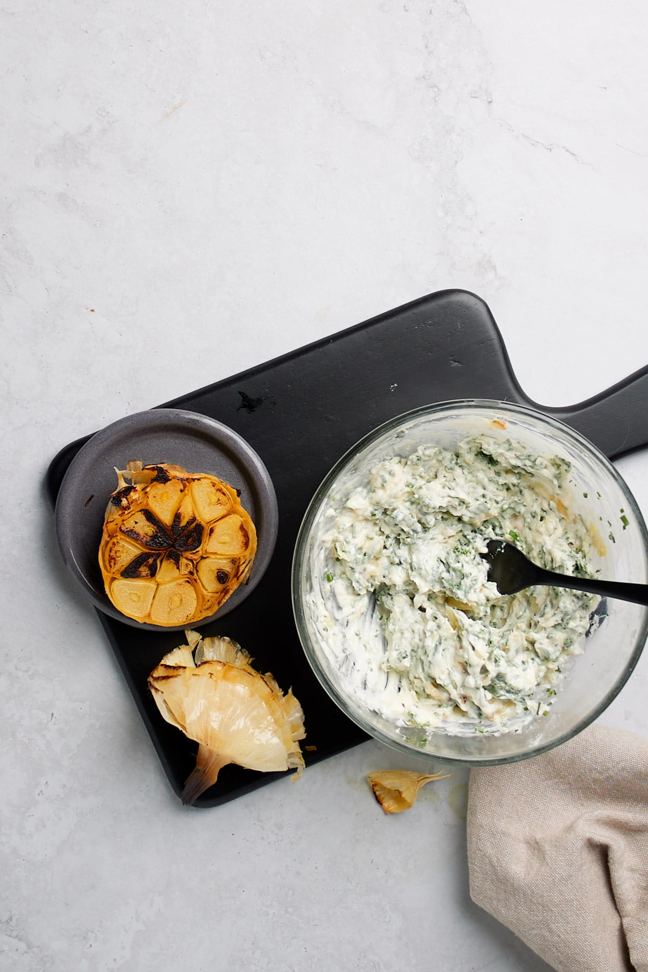 A bowl of creamy dip with green herbs sits on a black cutting board. Beside it, there is a halved, charred garlic bulb and a piece of what appears to be yellow onion skin. Featuring ingredients similar to those in our sheet pan gnocchi recipe, this savory spread pairs perfectly with herbed butter.