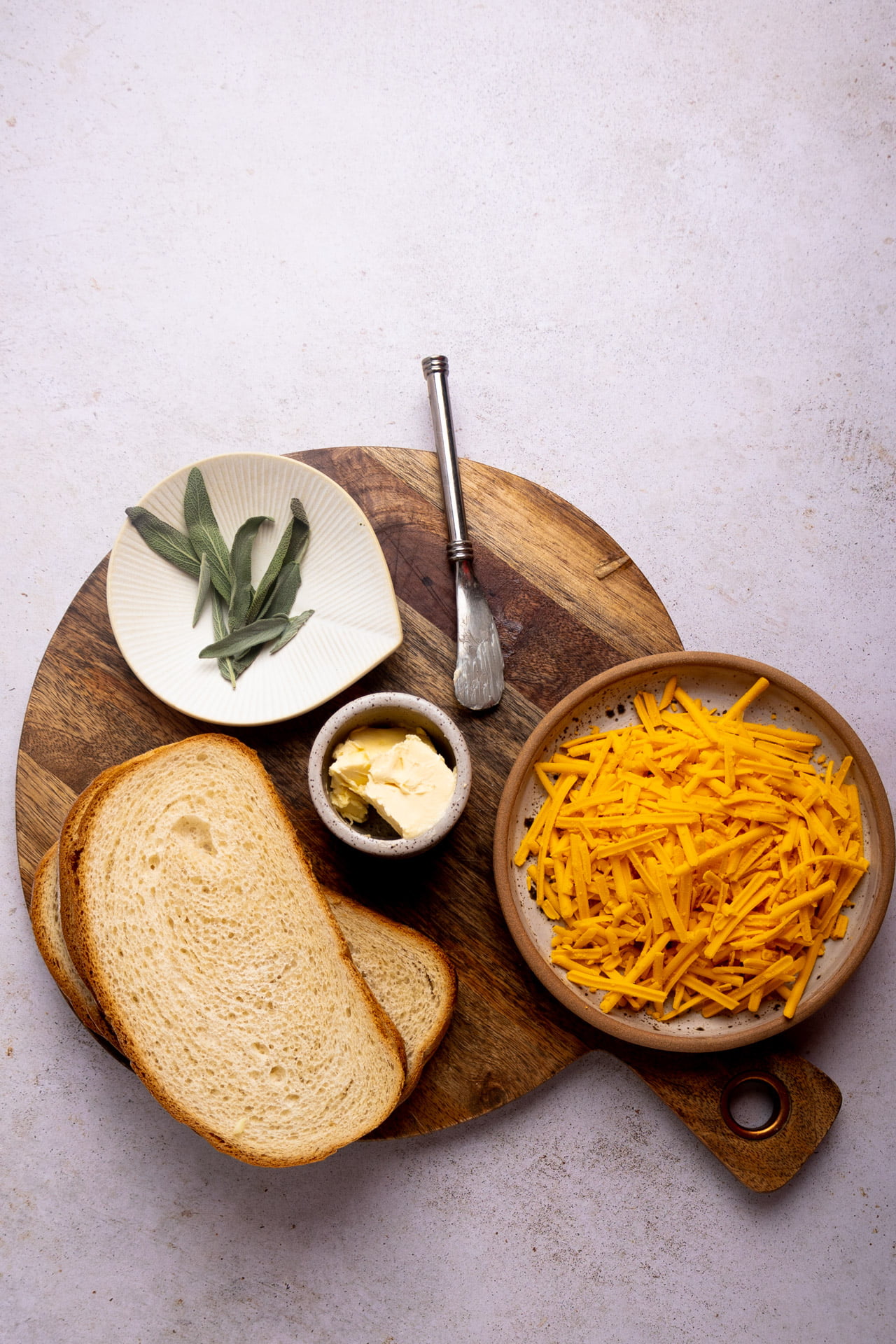 shredded cheddar, slices of bread and sage leaves on a round cutting board.