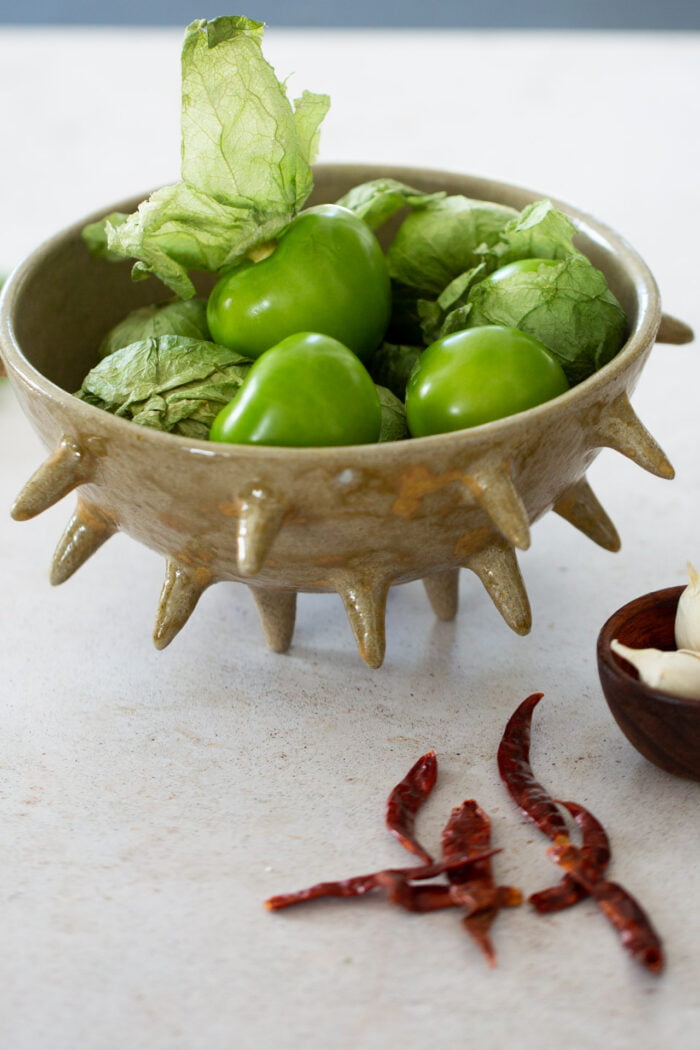 tomatillos in a spiky bowl, chile de arbol on the countertop and garlic in a small bowl.
