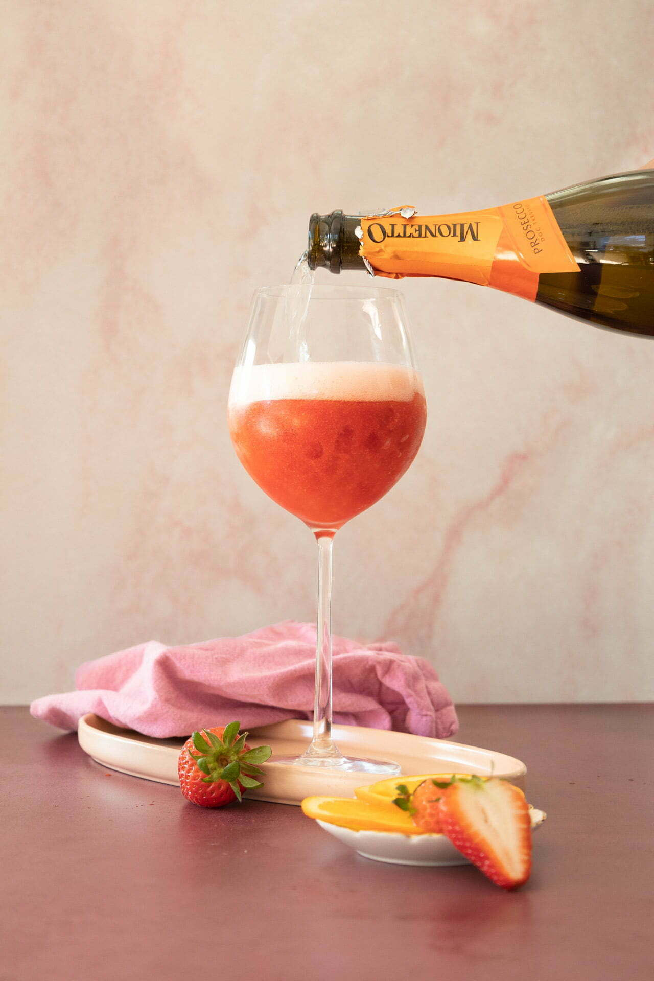 A glass of red-orange bubbly cocktail is being poured from a bottle of Martini Asti. The glass is filled halfway with frothy liquid. A fresh strawberry and slice of orange garnish the base, arranged on a wooden board with a pink cloth in the background against a mottled light backdrop, perfect for an Aperol spritz.
