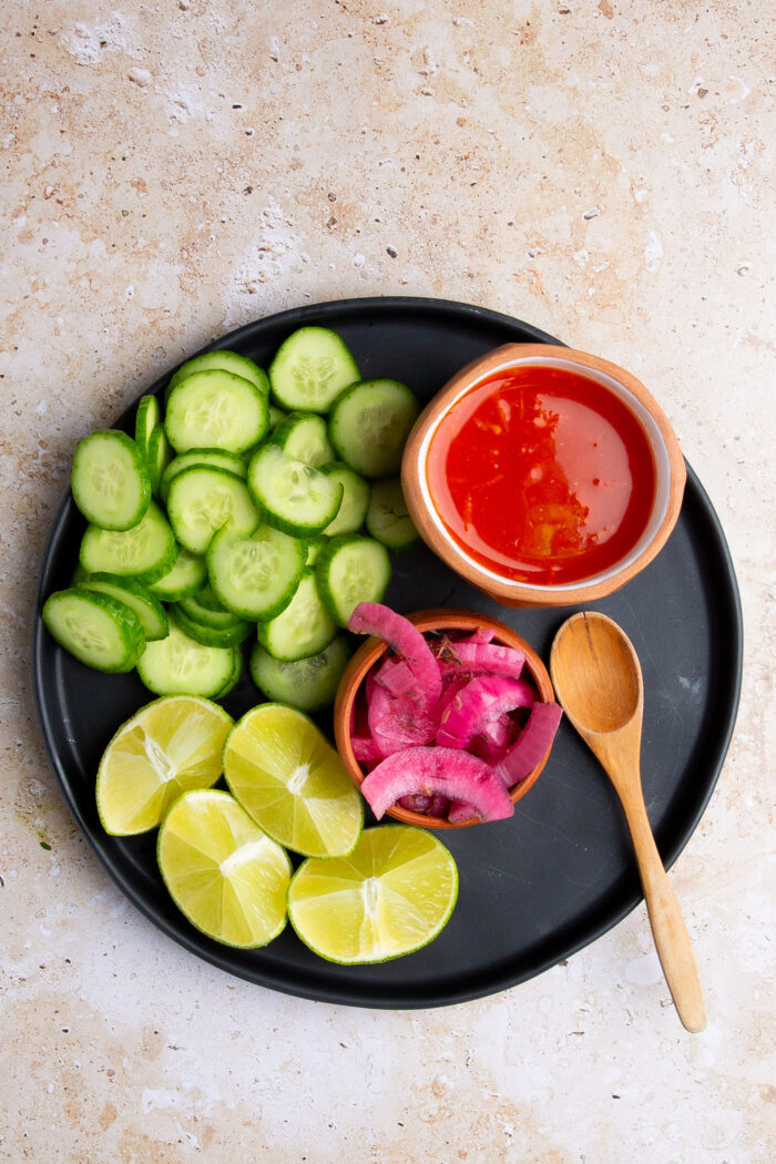 sliced cucumber, halved limes, salsa roja and Mexican pickled onions on black plate