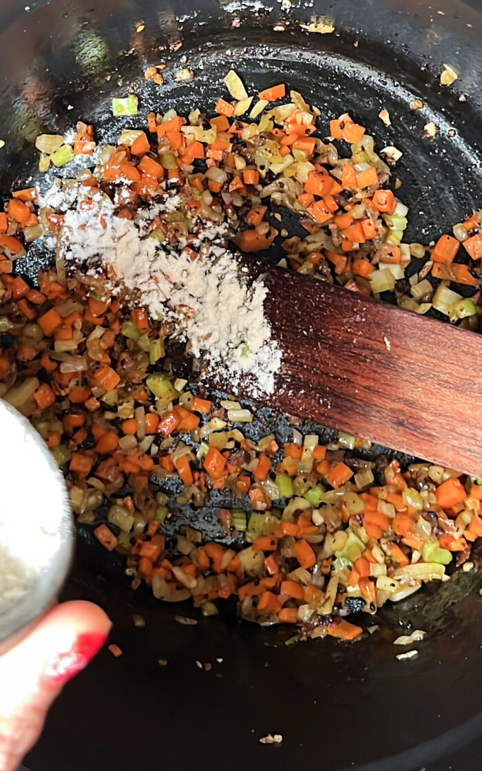 A person sprinkling flour over vegetables in a pan for a deliciously creamy soup.