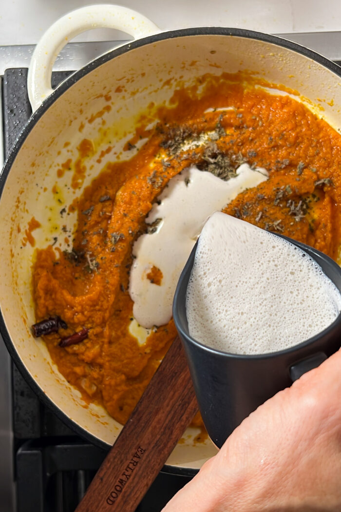 A person using a wooden spoon to stir a creamy vegan pumpkin sauce in a pan.