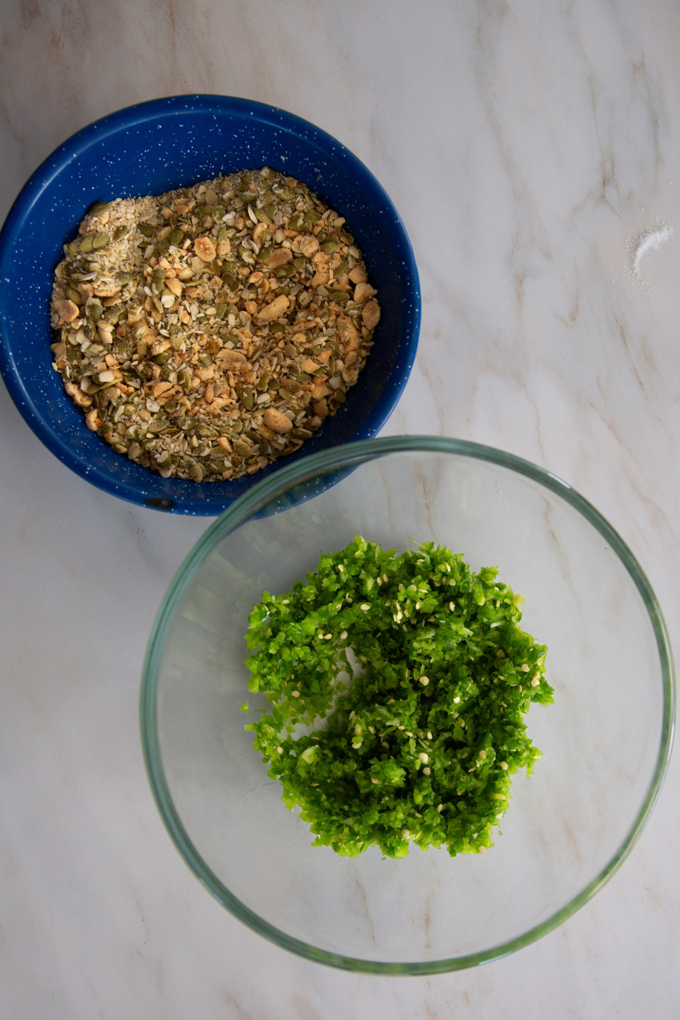 A top-down view of two bowls on a countertop containing chopped nuts and Serrano peppers respectively.