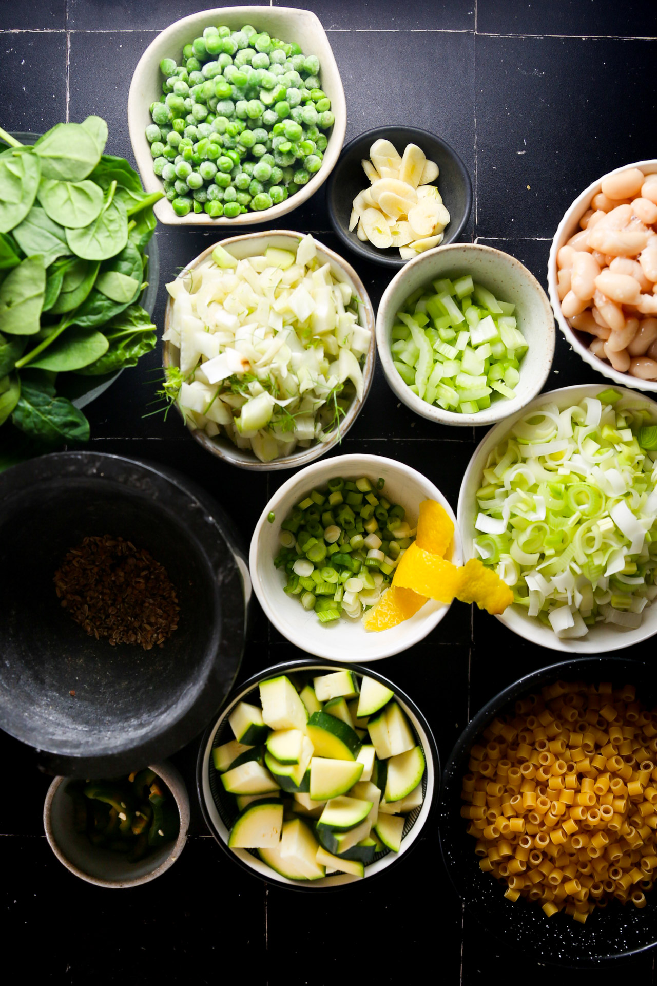 A variety of fresh ingredients carefully arranged in bowls on a black surface, including peas, spinach, sliced zucchinis, leeks, pasta, and spices, focusing on the colors and textures for preparation.