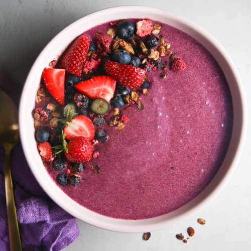 A creamy purple Smoothie Bowl is topped with fresh strawberries, blueberries, granola, and a few dried raspberries, arranged in clusters on the left side of the bowl. A gold spoon and a purple cloth napkin are placed to the left of the bowl on a light gray surface, making for an easy 5 Minute Recipe.