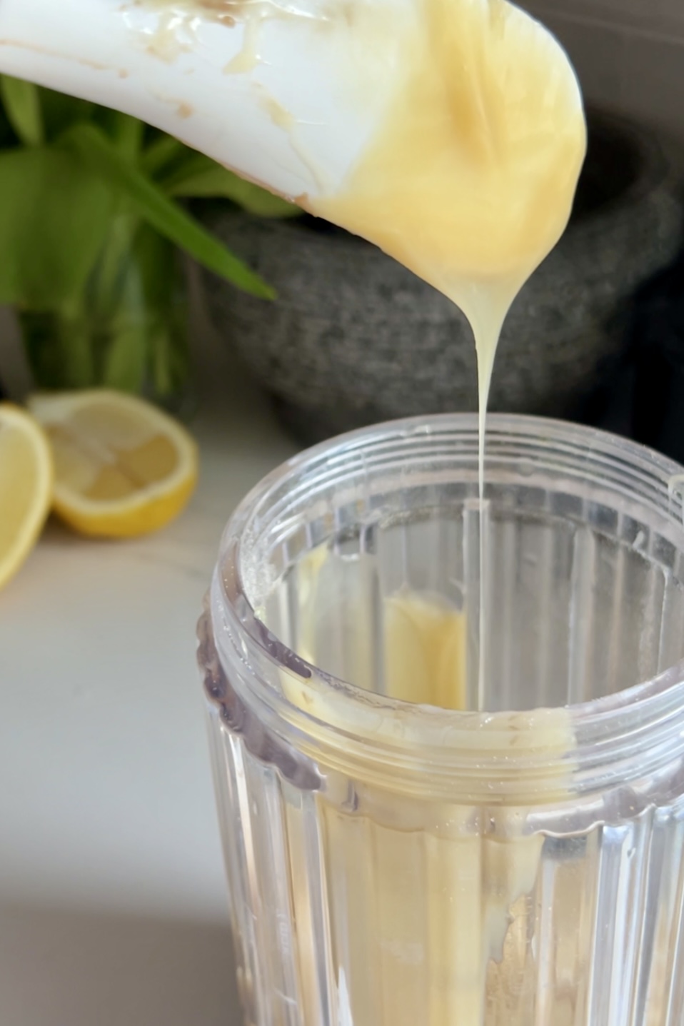 Condensed milk being poured from a white spoon into a clear glass, with a halved lemon and a stone mortar in the background on a kitchen counter for Lemon Pie Paletas recipe.