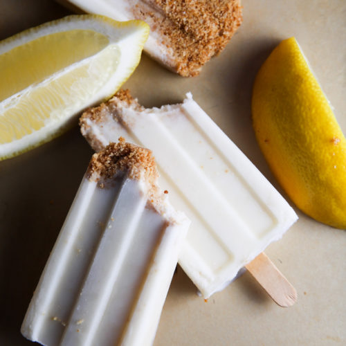 Three lemon pie-flavored paletas with brown sugar crust on a beige surface, accompanied by fresh lemon wedges. The paletas are partially visible and rest vertically.