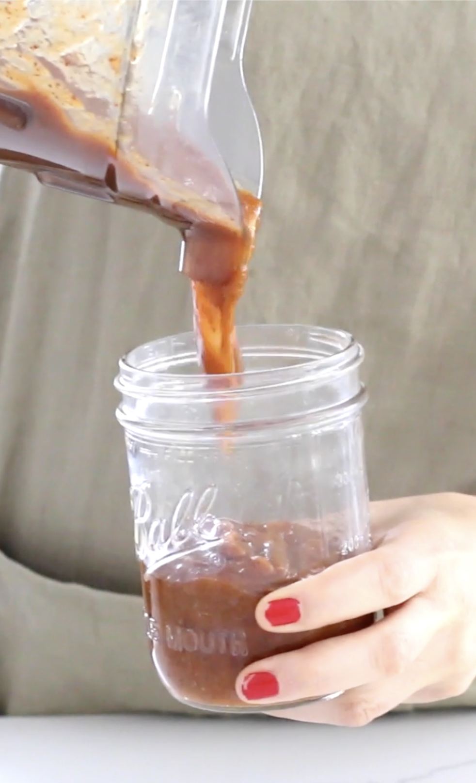 A person with red nail polish is pouring a thick, dark brown chamoy sauce from a clear blender into a glass mason jar, both held by hands partially visible in the image.