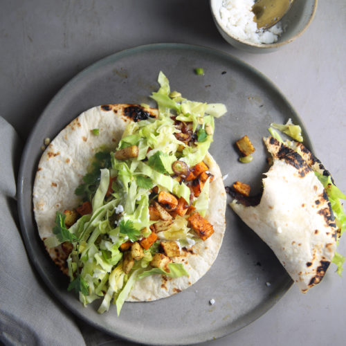 Two soft tacos filled with roasted vegetables, shredded lettuce, and crumbled cheese are placed on a gray ceramic plate. A small bowl with extra cheese and a golden spoon sits nearby. A piece of folded gray cloth is partially seen under the plate on the muted gray surface. Try this healthy recipe for sweet potato tacos!