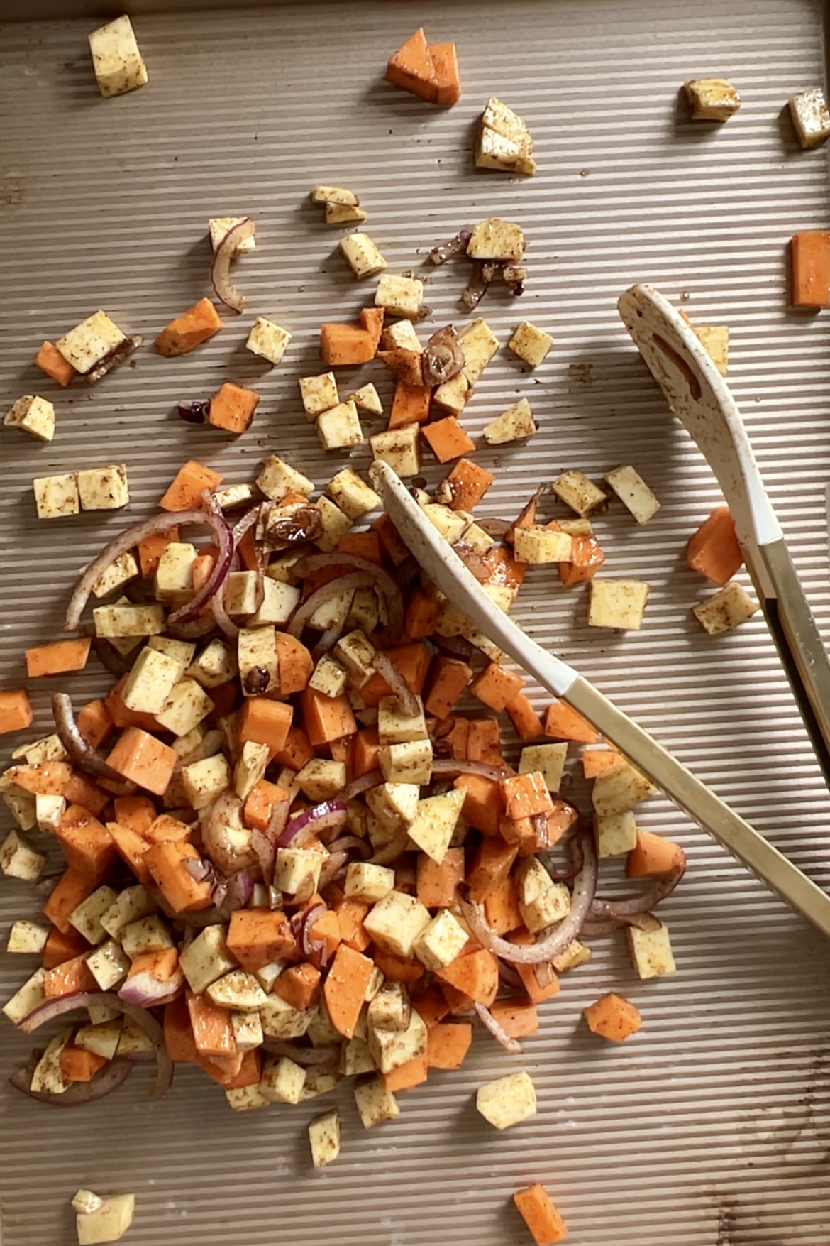 A baking sheet holds a healthy mixture of diced sweet potatoes, tofu cubes, and sliced onions. The ingredients are seasoned and spread out unevenly. Two white tongs are placed beside the mixture, lying across the baking sheet with ridges running horizontally—perfect for taco fillings.