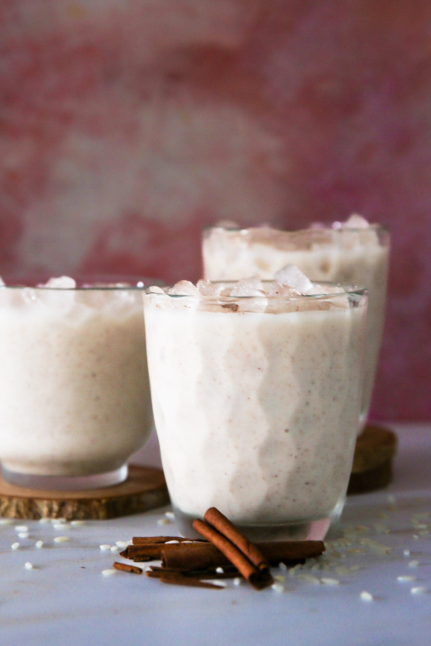 Three glasses of creamy vegan horchata topped with ice cubes, positioned on wooden coasters. The drinks are set against a soft pink background, garnished with cinnamon sticks and scattered rice grains in the