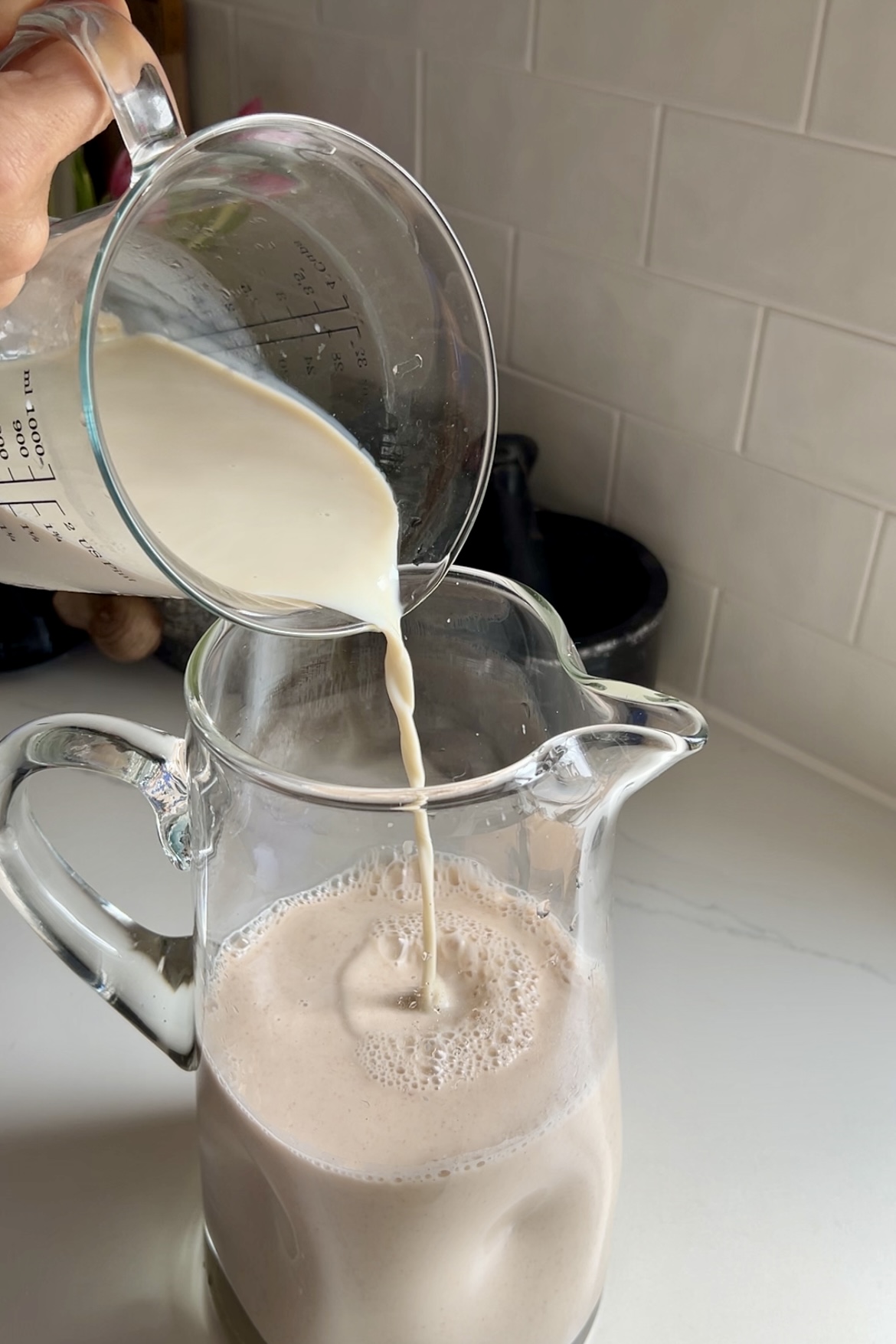 A person pours a stream of creamy milk from a measuring cup into a glass pitcher filled with frothy, light brown vegan horchata, set against a kitchen backsplash.