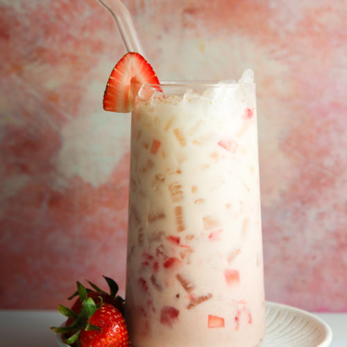 A refreshing strawberry horchata in a clear glass, filled with ice and diced strawberries, topped with a whole strawberry on the rim. The drink is served with a glass straw, on a light plate