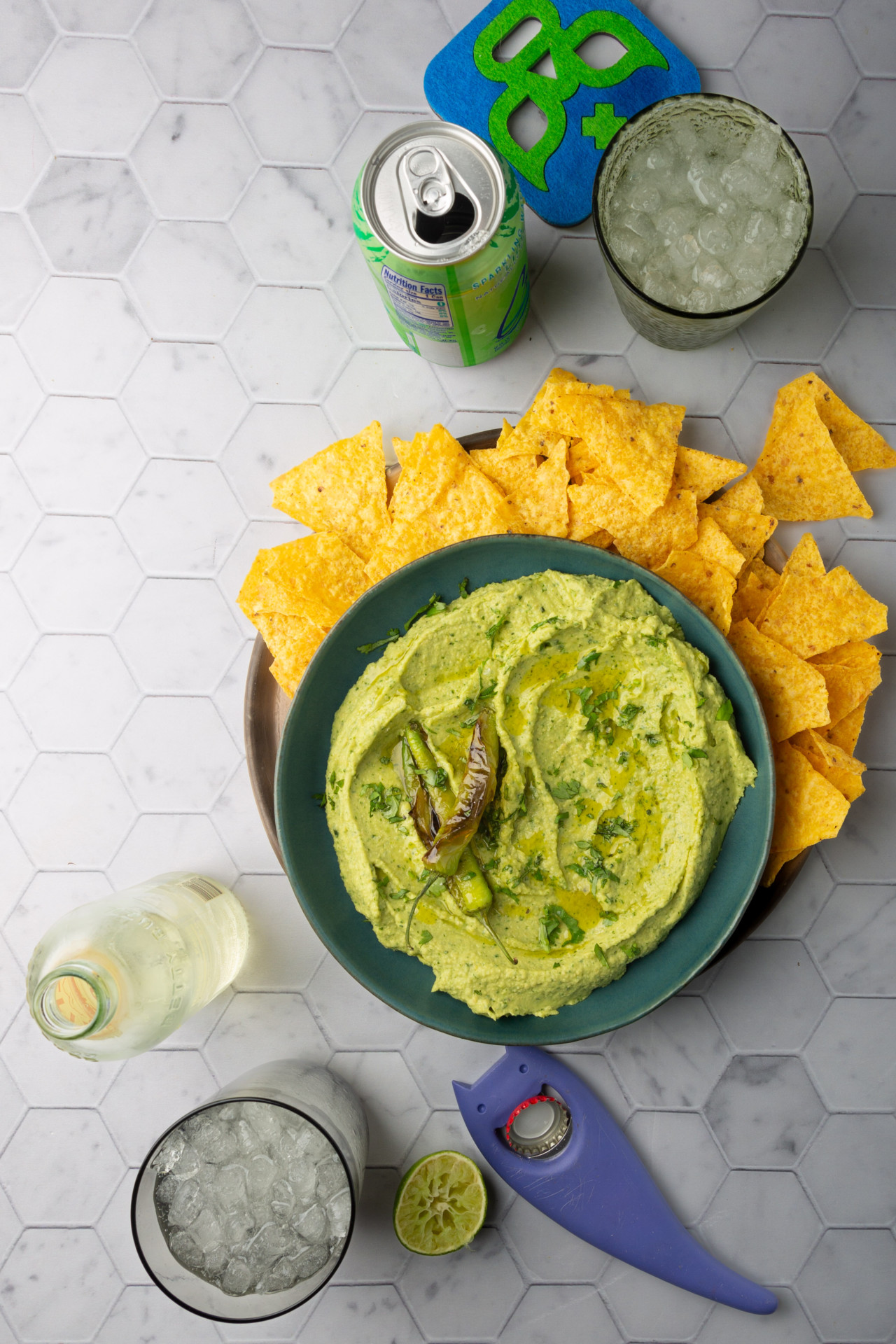 A bowl of avocado hummus topped with grilled jalapenos, surrounded by corn tortilla chips on a hexagon-patterned surface. Nearby are drinks, including a green can and a clear bottle, both served in glasses with ice. A juiced lime half and a blue shark-shaped bottle opener complete the scene.