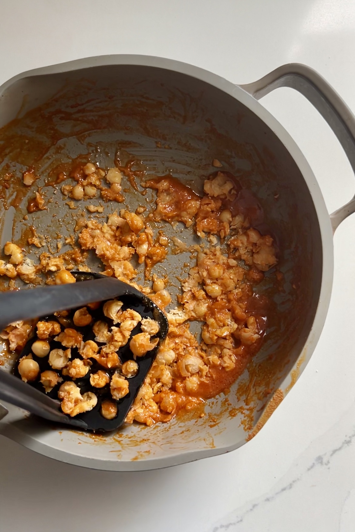 A non-stick saucepan containing smashed chickpeas mixed with a Buffalo red sauce. A black slotted spoon is positioned on the left side of the pan, partially mashing some chickpeas. The pan has two handles, one on each side. The background is a white surface.