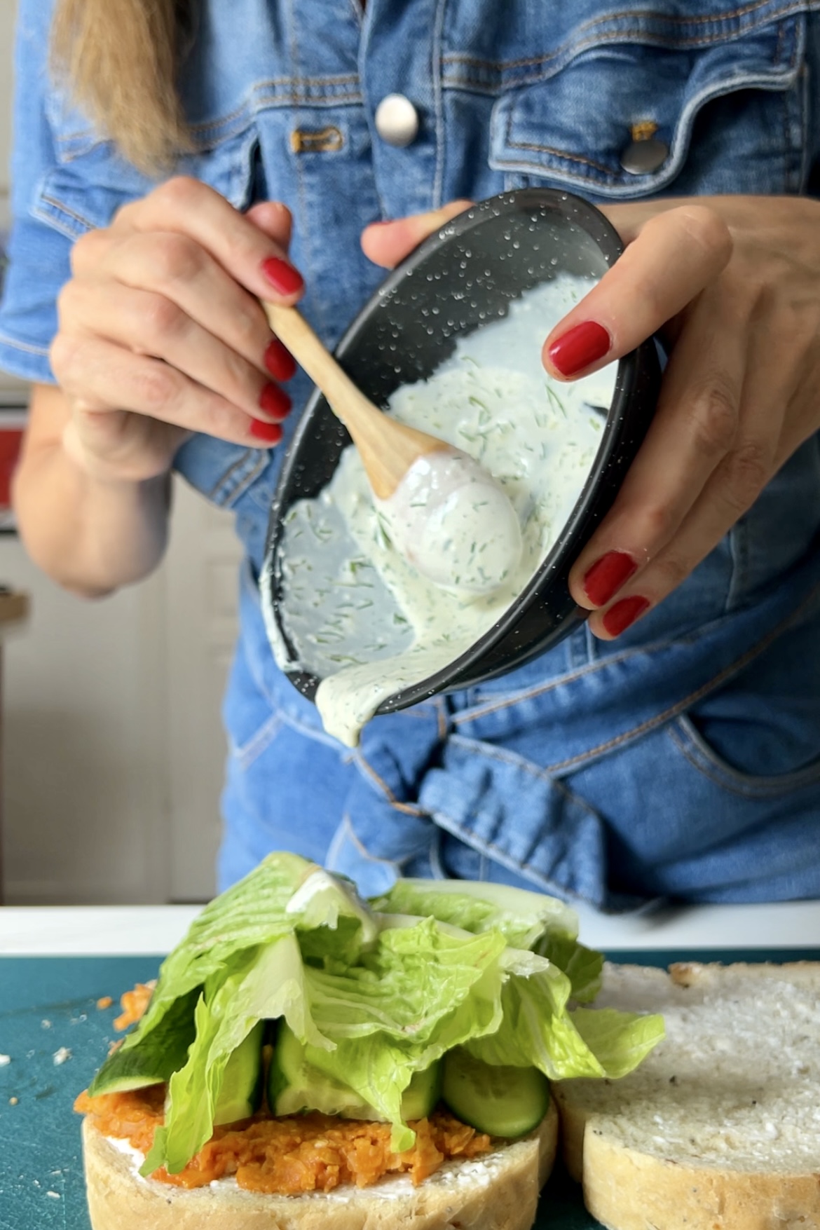 A person with red nail polish spreads creamy dressing from a black bowl onto a smashed chickpea and lettuce sandwich using a wooden spoon. The individual, clad in a blue denim shirt, stands by the kitchen counter, meticulously assembling their delicious meal.