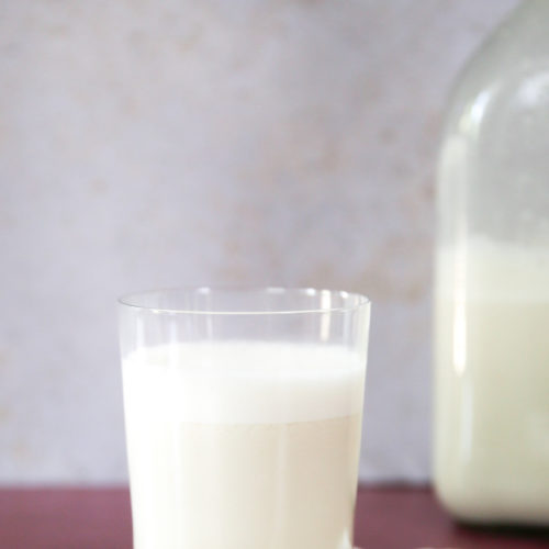 A clear glass filled with creamy cashew milk sits on a wooden coaster. Three cashew nuts are placed beside the glass. In the background, a large glass bottle containing more cashew milk is slightly out of focus. A white folded napkin lies under the glass, all on a brown surface. Make cashew milk using the easiest way and enjoy this delightful drink!