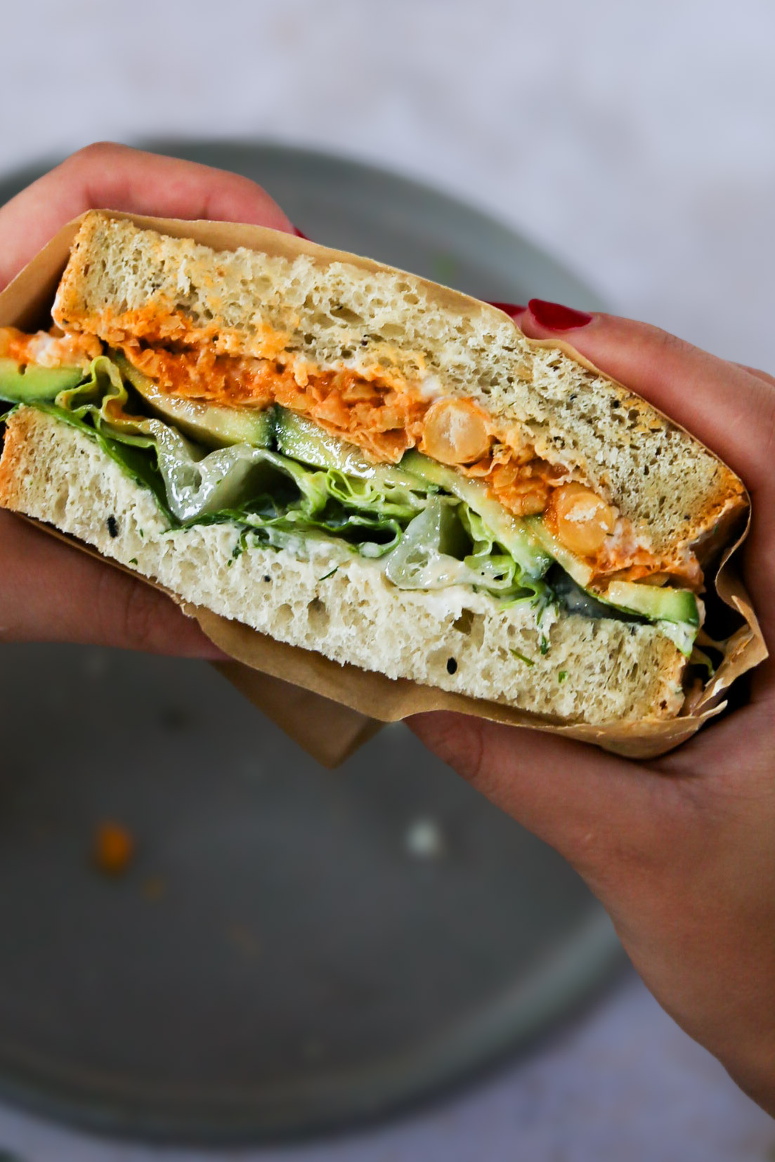 A close-up of a sandwich being held by two hands. The sandwich, featuring smashed chickpeas, consists of two thick slices of bread filled with leafy greens and cucumber slices. It is partially wrapped in brown paper. The background is a blurred, gray surface.
