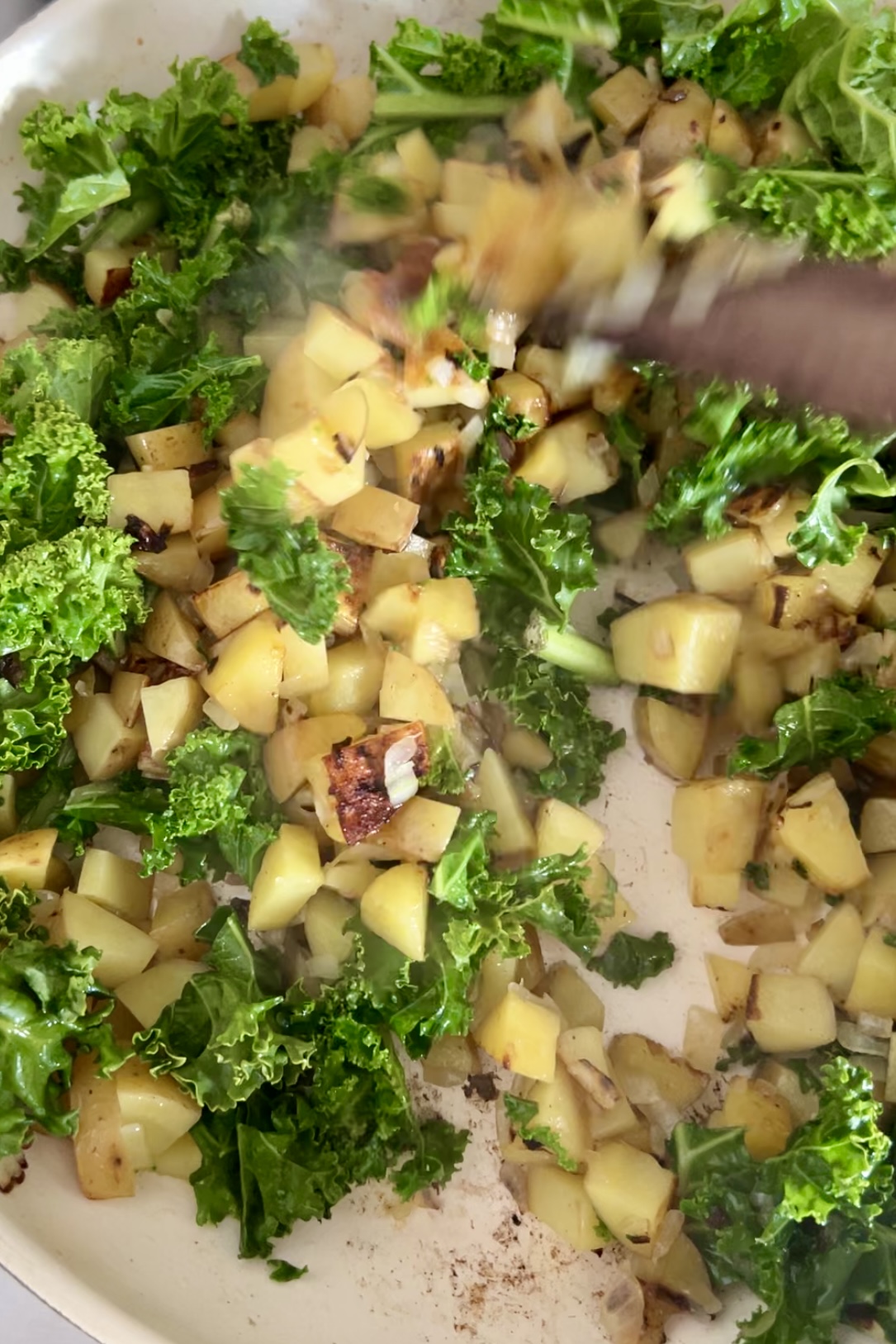A close-up image shows a pan filled with diced potatoes and fresh kale being stirred. The potatoes are lightly browned, indicating they have been sautéed or fried. The greens look fresh and vibrant, contrasting with the golden-brown cubes of potato. Steam rises from the hot pan, perfect for Kale Potato Tacos.