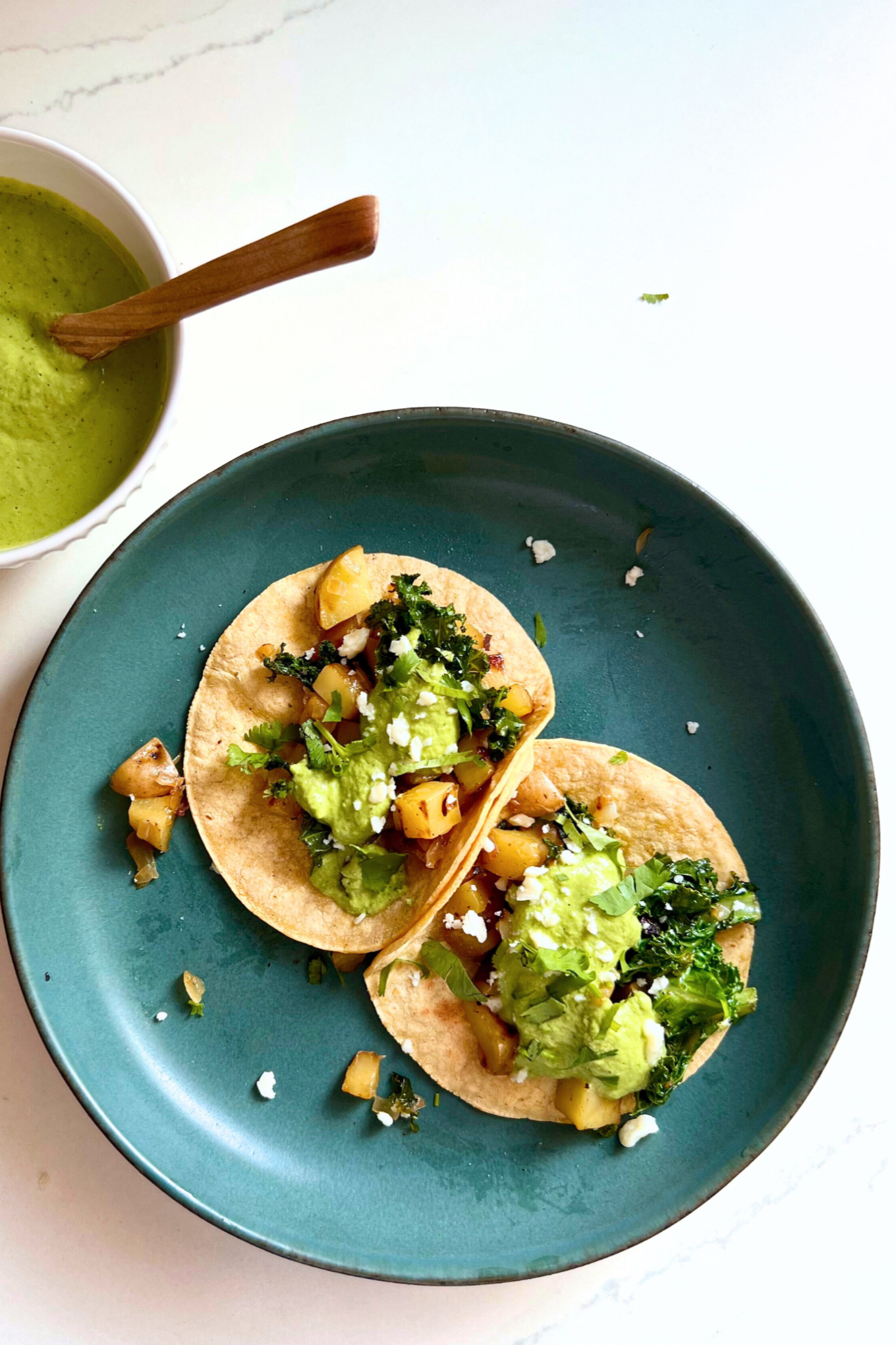 Two soft tacos filled with sautéed vegetables, kale, and crumbled cheese sit on a blue-green plate. Nearby, a bowl of cilantro Bitchin Sauce with a wooden spoon is placed on a white surface. Small bits of taco filling and cheese crumbs surround the tacos on the plate.