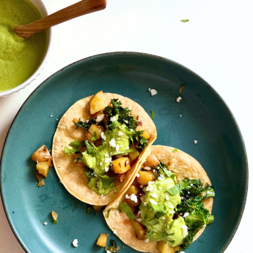 Two soft tacos filled with sautéed vegetables, kale, and crumbled cheese sit on a blue-green plate. Nearby, a bowl of cilantro Bitchin Sauce with a wooden spoon is placed on a white surface. Small bits of taco filling and cheese crumbs surround the tacos on the plate.